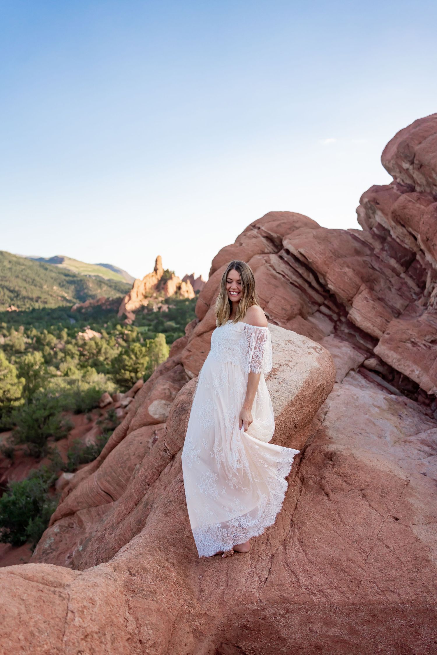 White lace maternity dress flows in the wind atop red rock formations in nature.