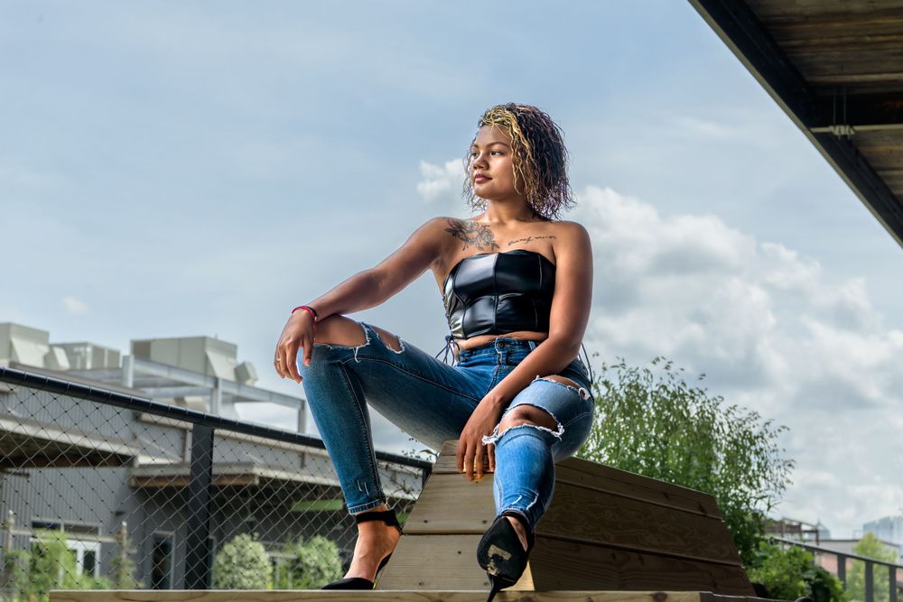 A person in ripped jeans and a black crop top sits casually on an outdoor structure against a cloudy blue sky.