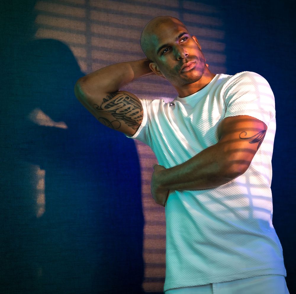 Athletic person in white t-shirt with tattoos poses against dramatic blue lighting and window shadows.