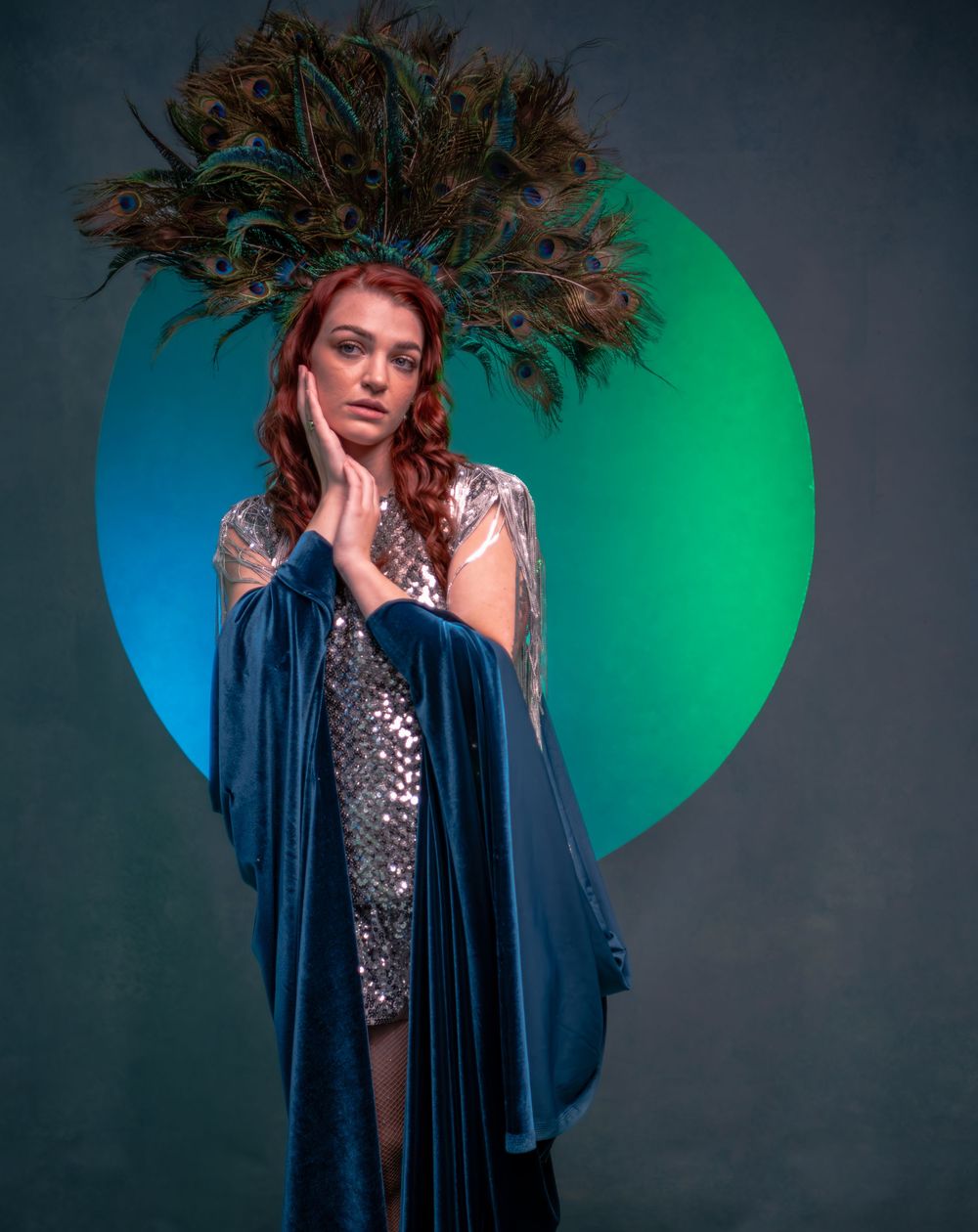 A figure in a dark blue velvet cape poses with peacock feathers against a vibrant green and blue circular backdrop.