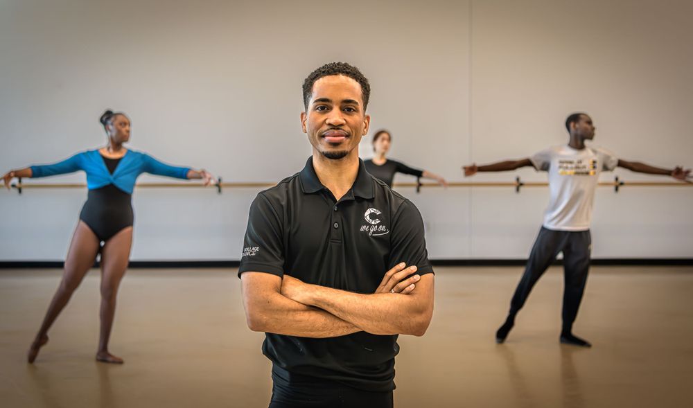 Dance instructor stands with crossed arms in studio as dancers practice poses in the background.