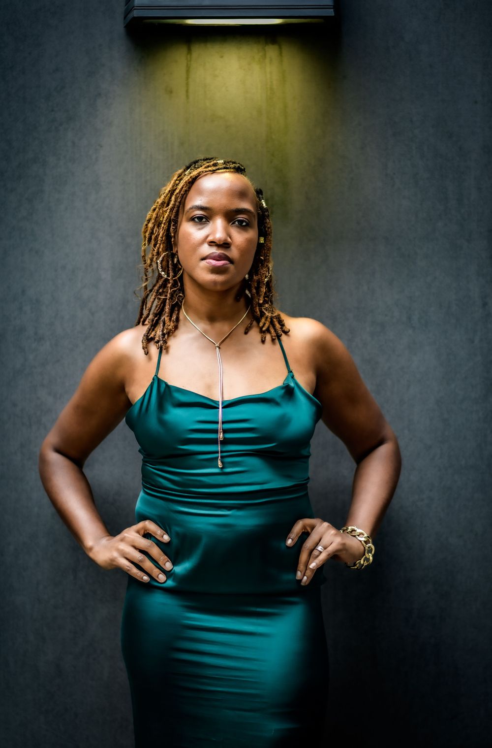 Woman in emerald green satin dress poses against dark wall with dramatic overhead lighting.
