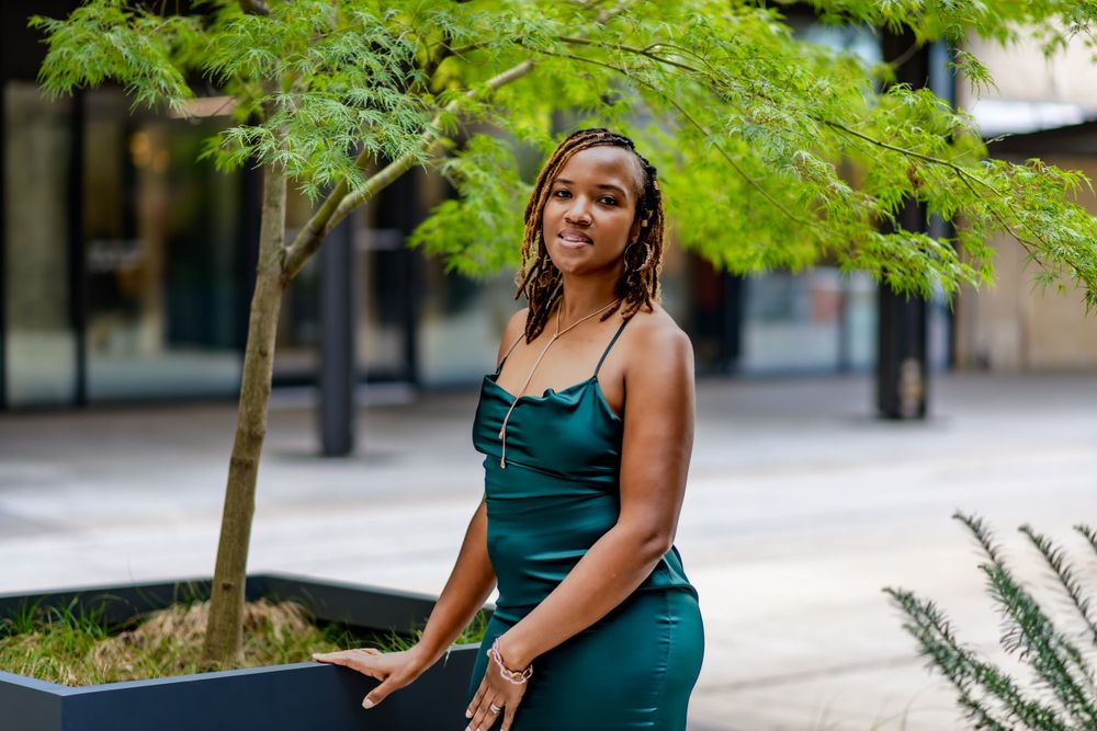 Someone in an elegant dark green dress sits next to a decorative outdoor planter with a small tree in an urban setting.