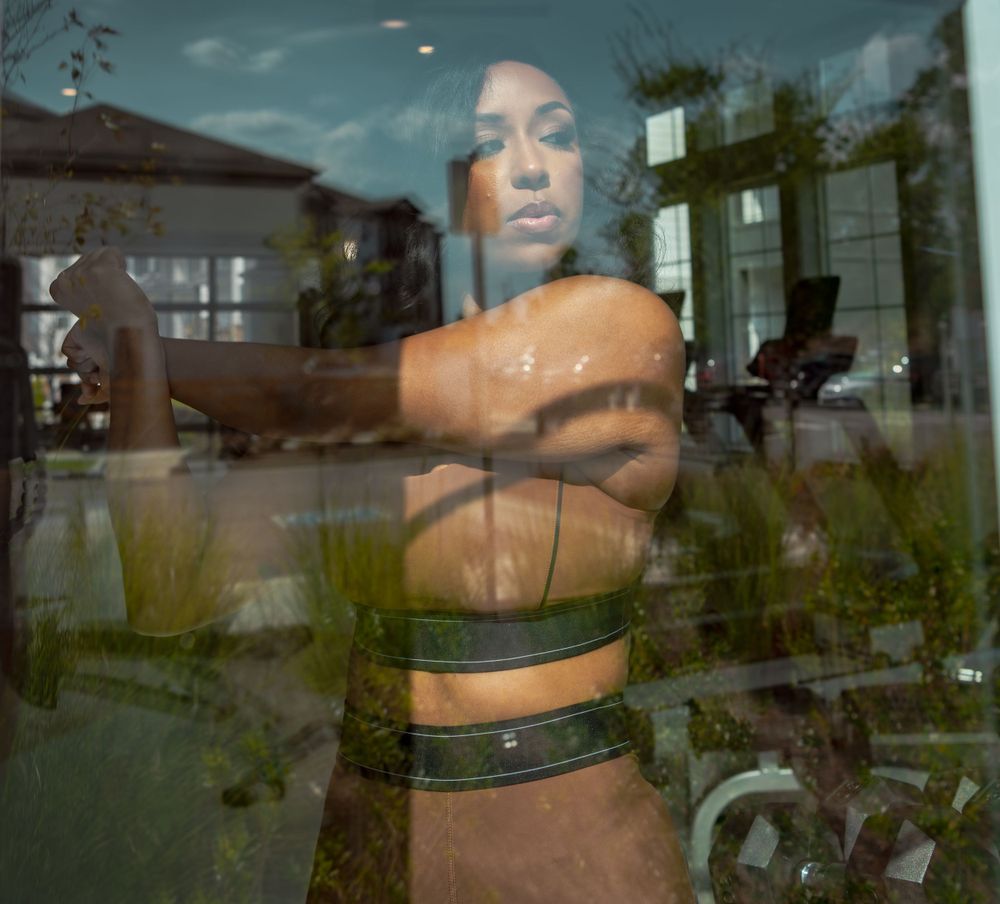 Model in undergarments displayed in a window with reflections and greenery visible outside.