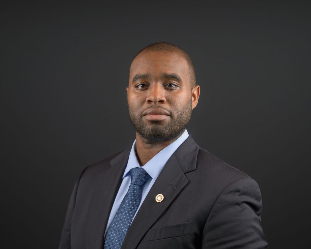 Professional headshot of a person wearing a dark suit with blue tie against a black background.
