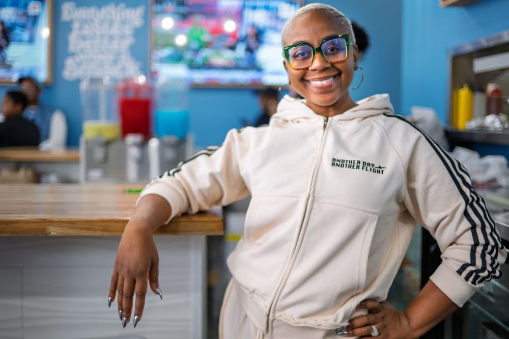 A smiling person wearing glasses and a cream-colored hoodie stands casually in a cafe with blue walls.
