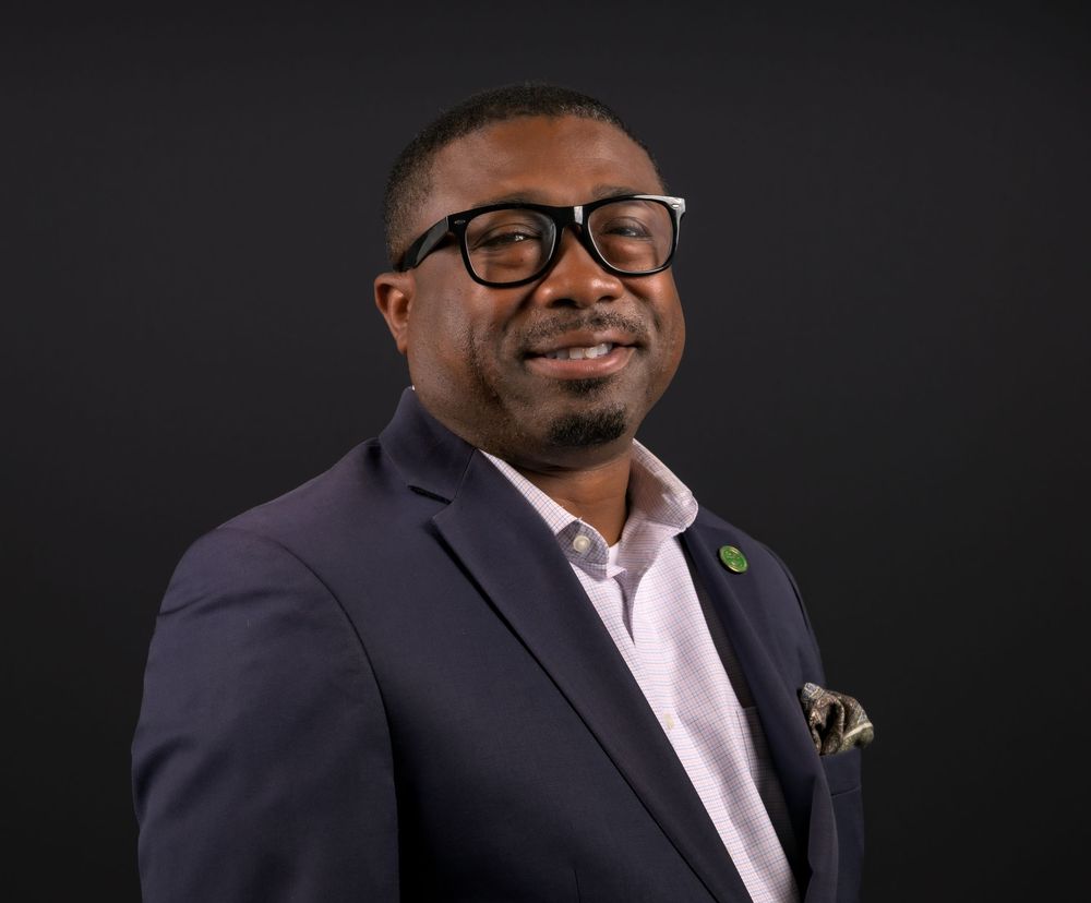 Professional headshot series showing someone in a dark suit and glasses against a black background, smiling warmly.