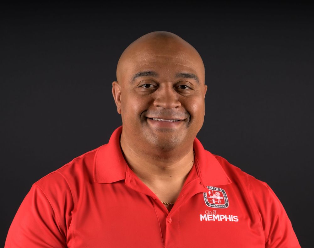 A person in a red polo shirt smiles warmly against a black background in a professional headshot series.