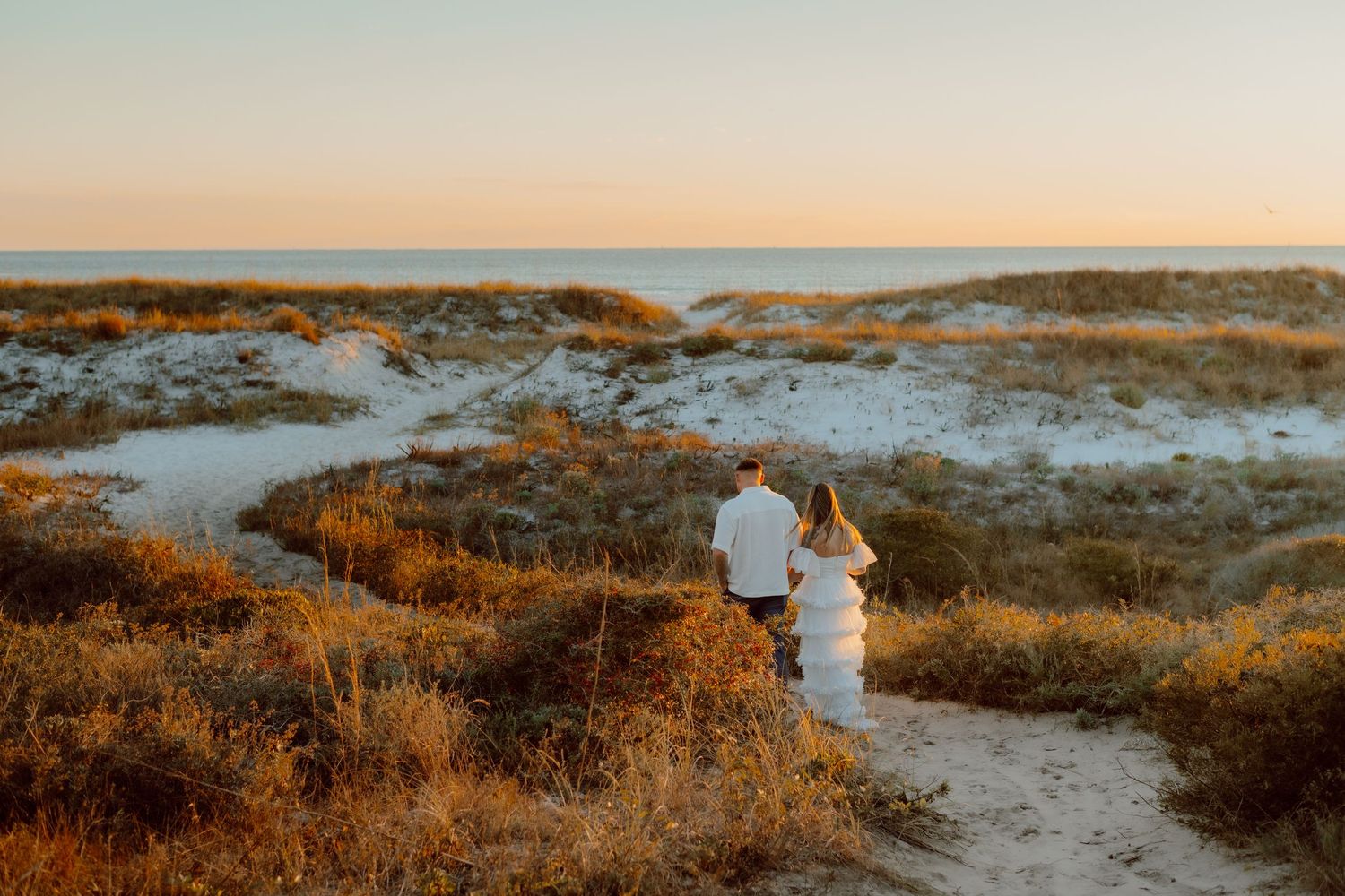Couple in wedding attire holding hands while walking along beach with crashing waves.