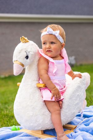 Baby wearing pink rides on plush white swan toy outdoors on blue blanket.