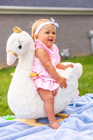 Baby in pink outfit sitting with plush white swan rocker toy on blue blanket outdoors.