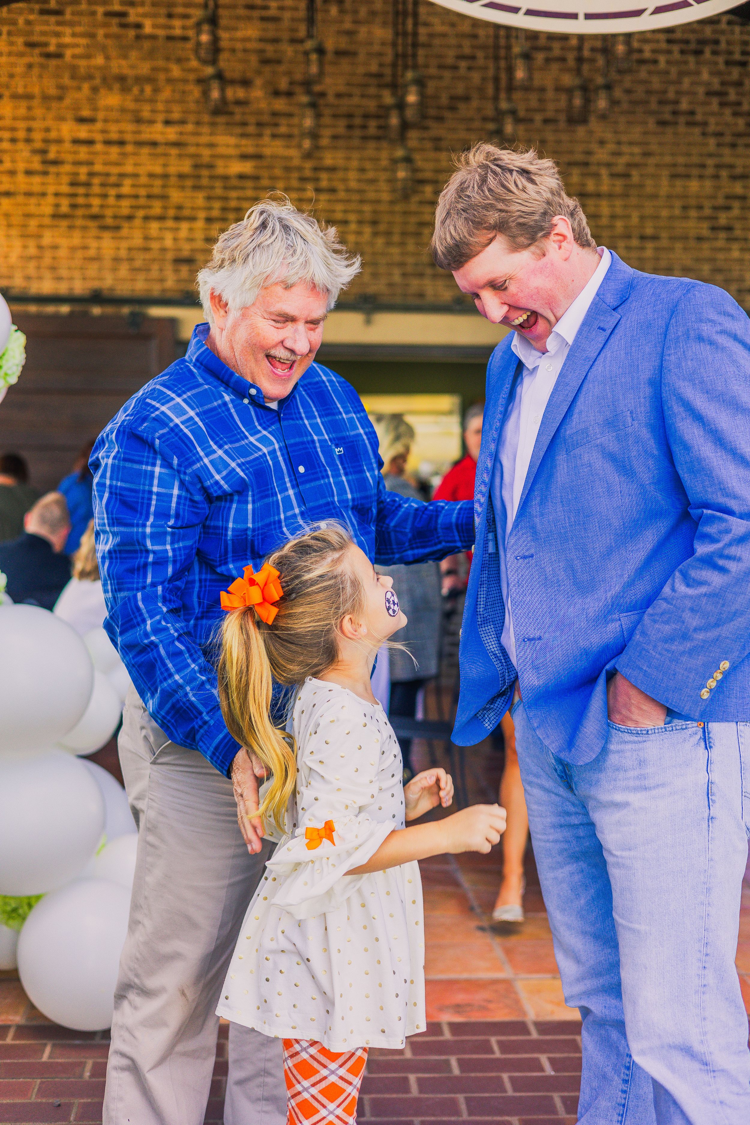 Family members sharing a sweet dance moment at outdoor event with brick backdrop and white balloons.