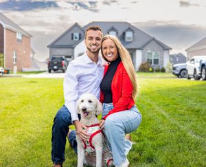 Family poses with their white fluffy dog wearing red bandana on sunny lawn.