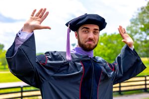Graduate in black cap and gown raises arms in celebratory poses against outdoor backdrop.