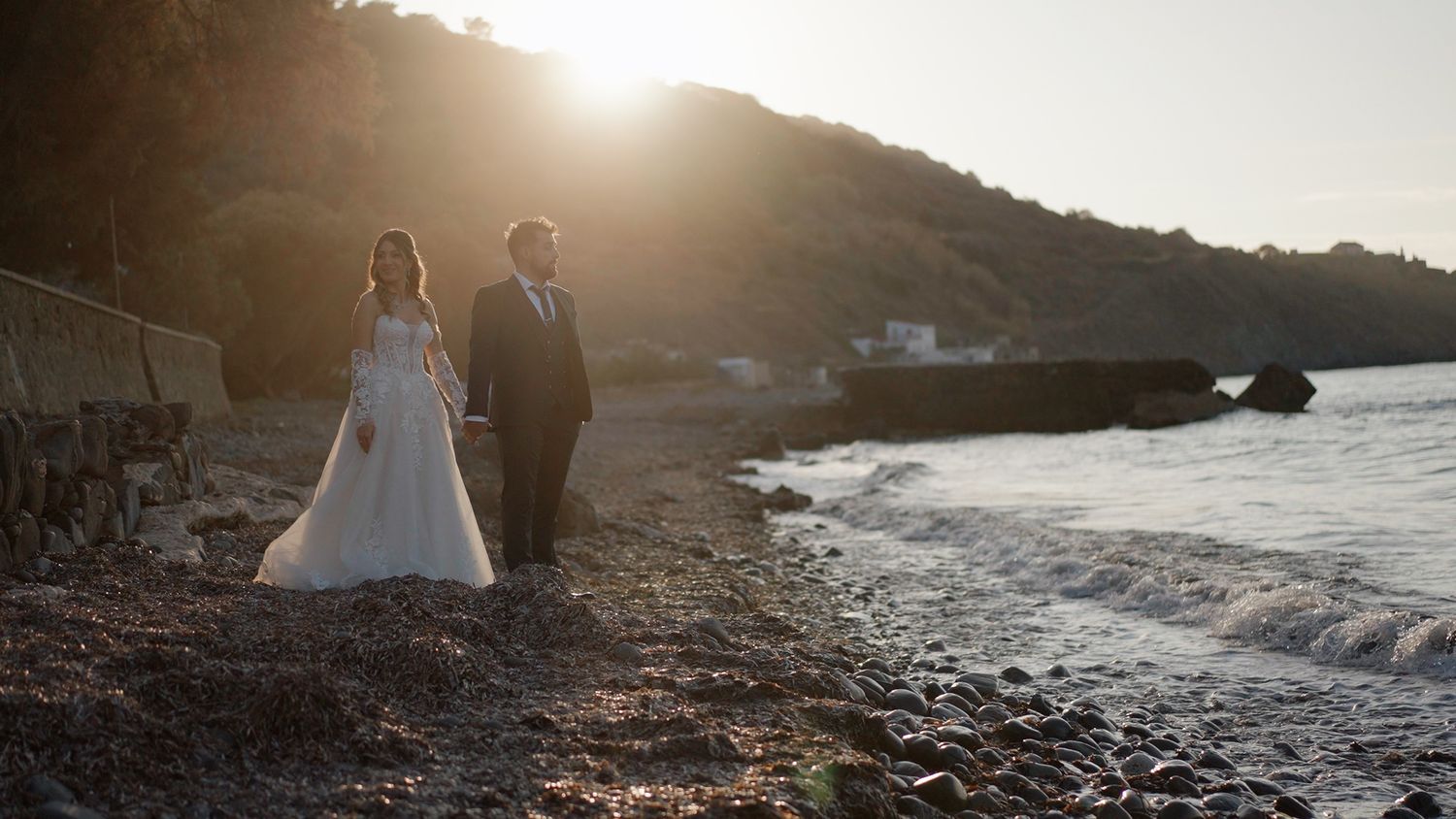 A couple walks together on a rugged coastal hillside at golden hour with the ocean visible in the distance.