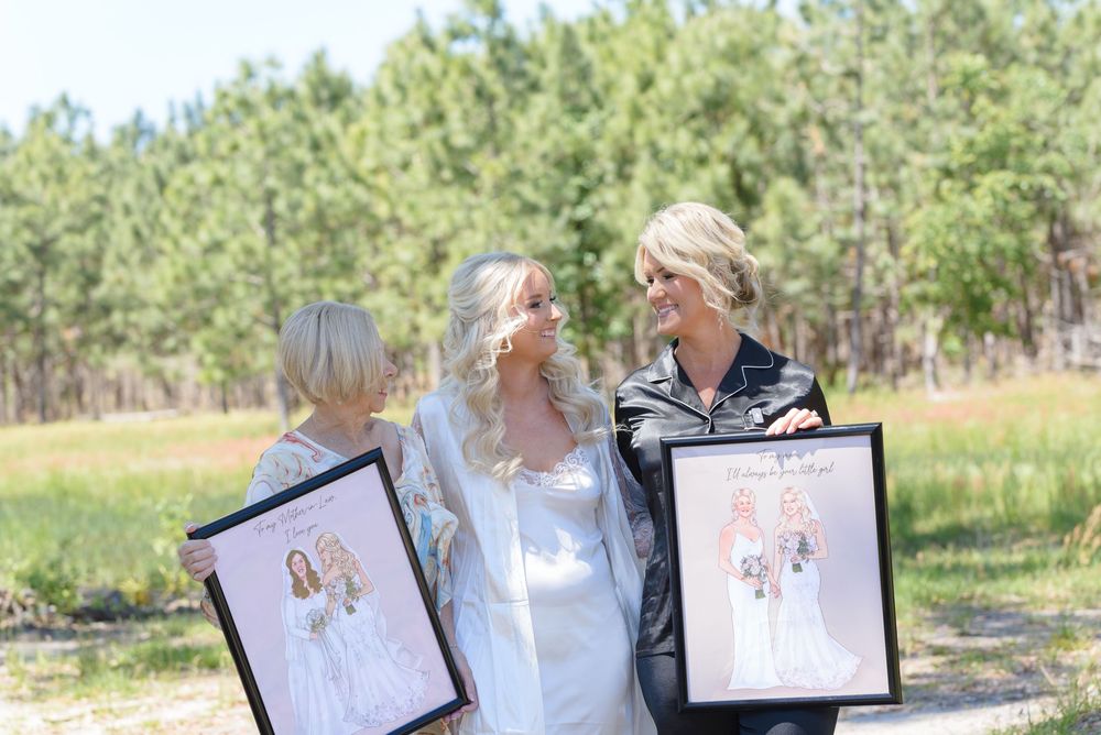 Three people hold framed artwork while laughing together outdoors in a sunny field with pine trees in the background.