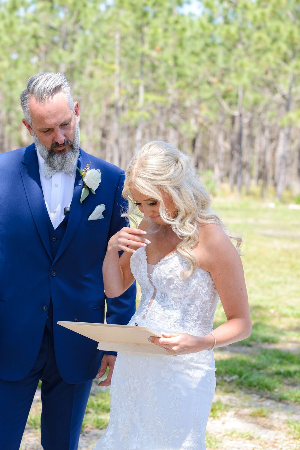 Bride in lace dress reacts emotionally while reading letter outdoors next to person in navy suit.