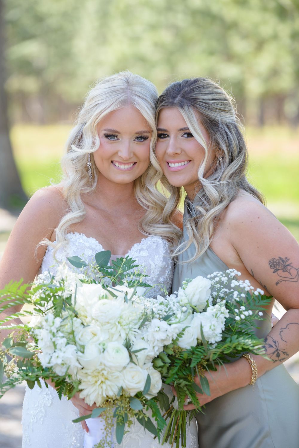 A bride and bridesmaid stand together smiling while holding a lush white floral bouquet with greenery outdoors.