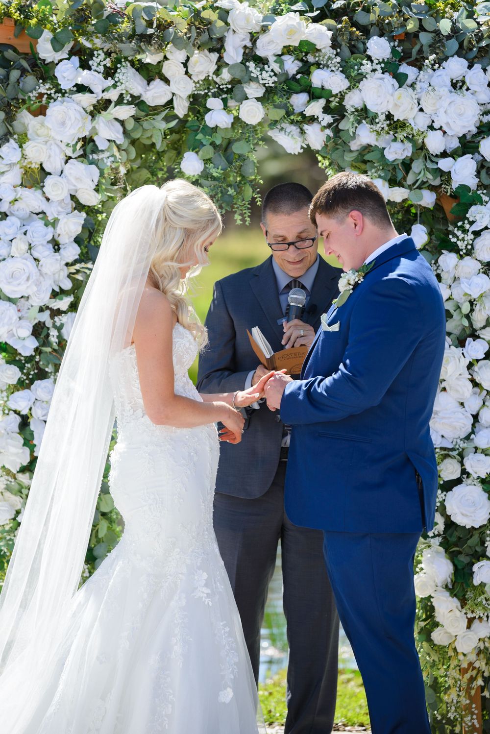 A wedding ceremony under a white floral arch with a couple exchanging rings as an officiant presides over the moment. little red wagon farms