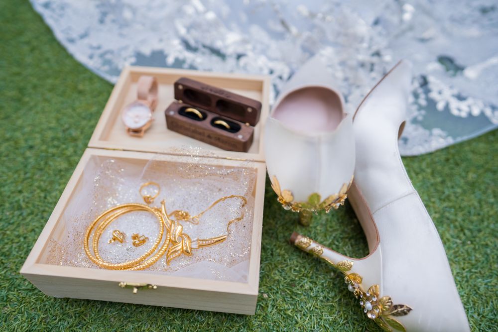 Wedding accessories including rings in a box, a white high heel shoe, and jewelry laid out on grass.