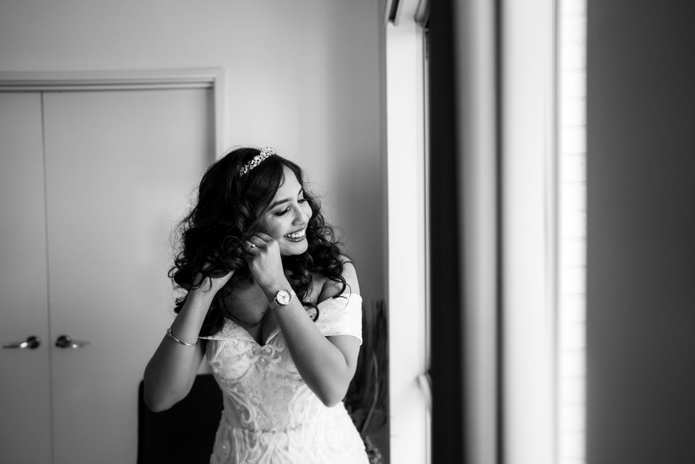 Black and white photo of someone in a wedding dress putting on earrings near a window.