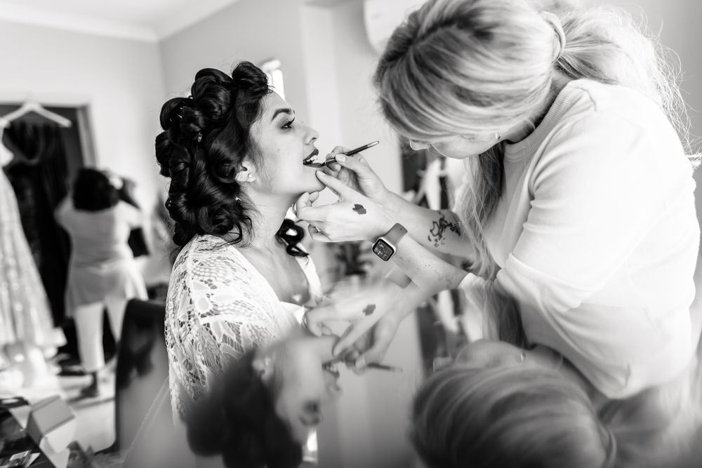 Black and white photo of wedding preparation with makeup being applied in a bright room.