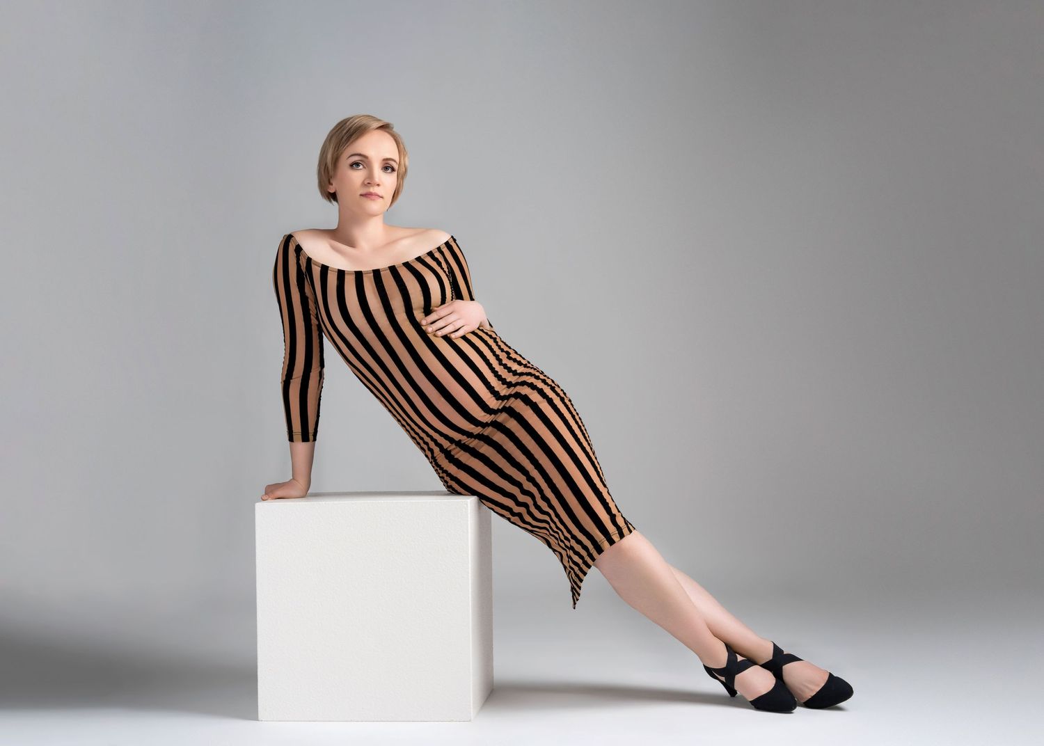 a pregnant woman wearing a skin tight striped dress leans on a white cube during a maternity photo shoot in studio
