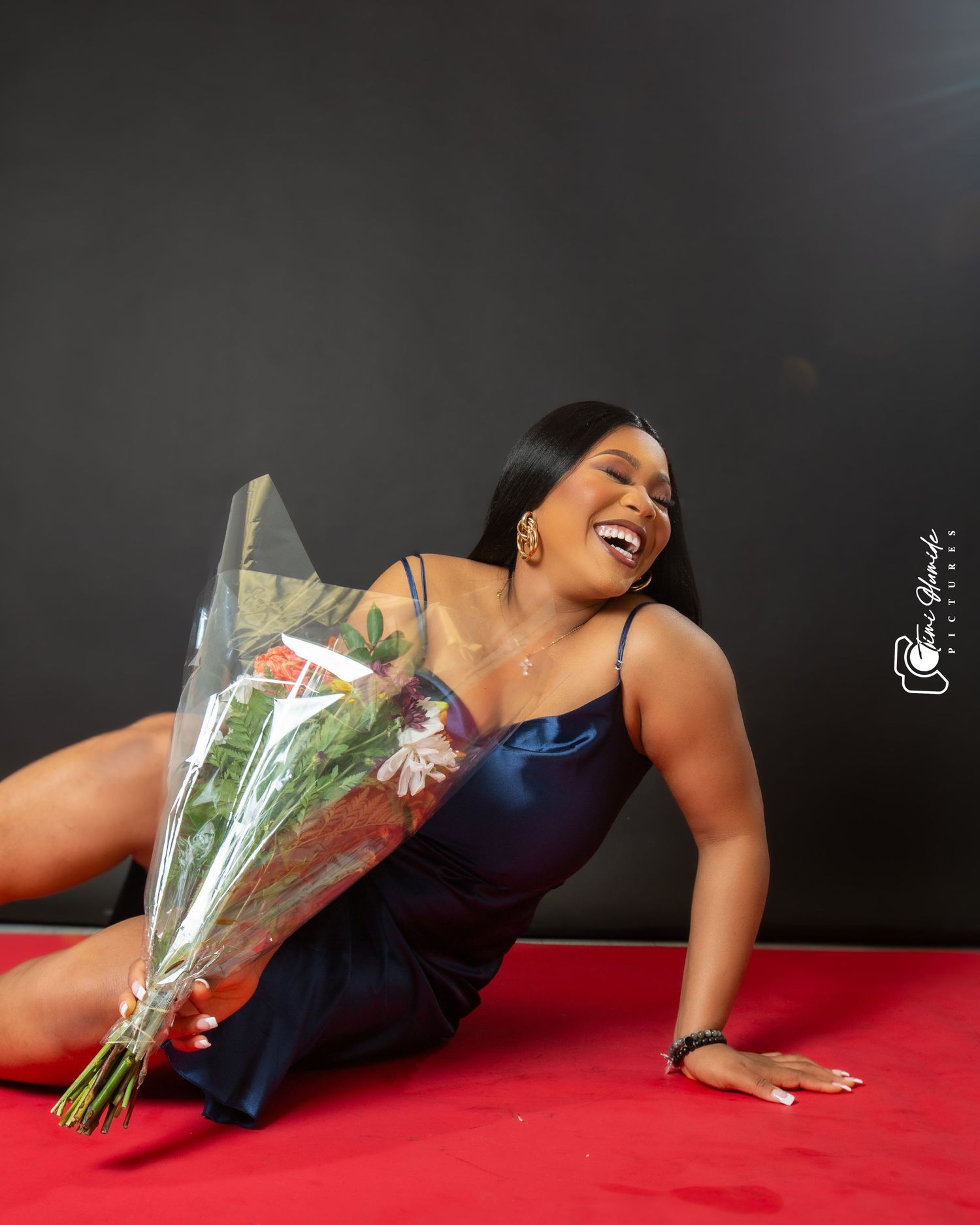 Joyful model in navy dress lies on red surface holding bouquet of flowers.
