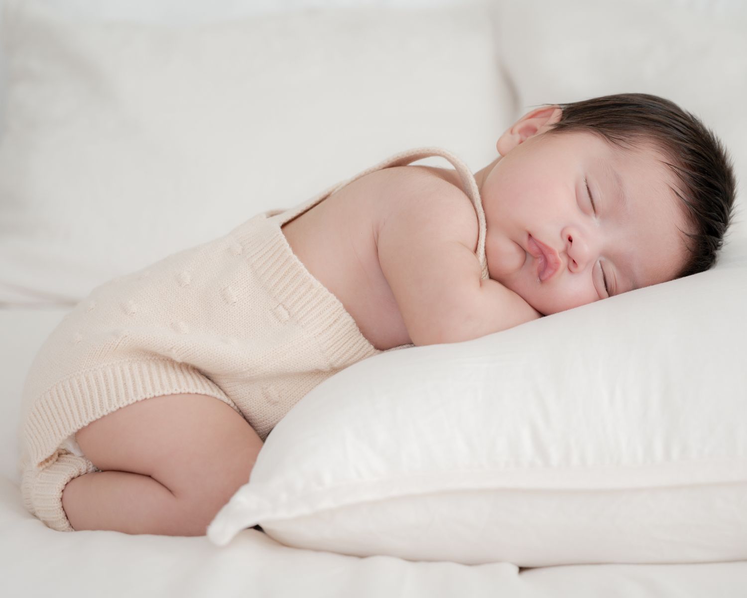 Sleeping infant in cream-colored outfit resting on white pillow.