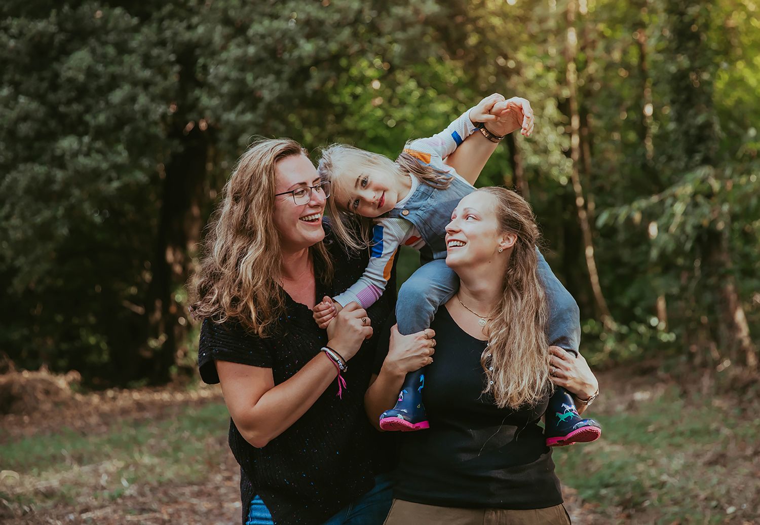 Two people enjoy a playful moment outdoors with a small child on a sunny autumn day in a wooded setting. During a photoshoot in Bexley