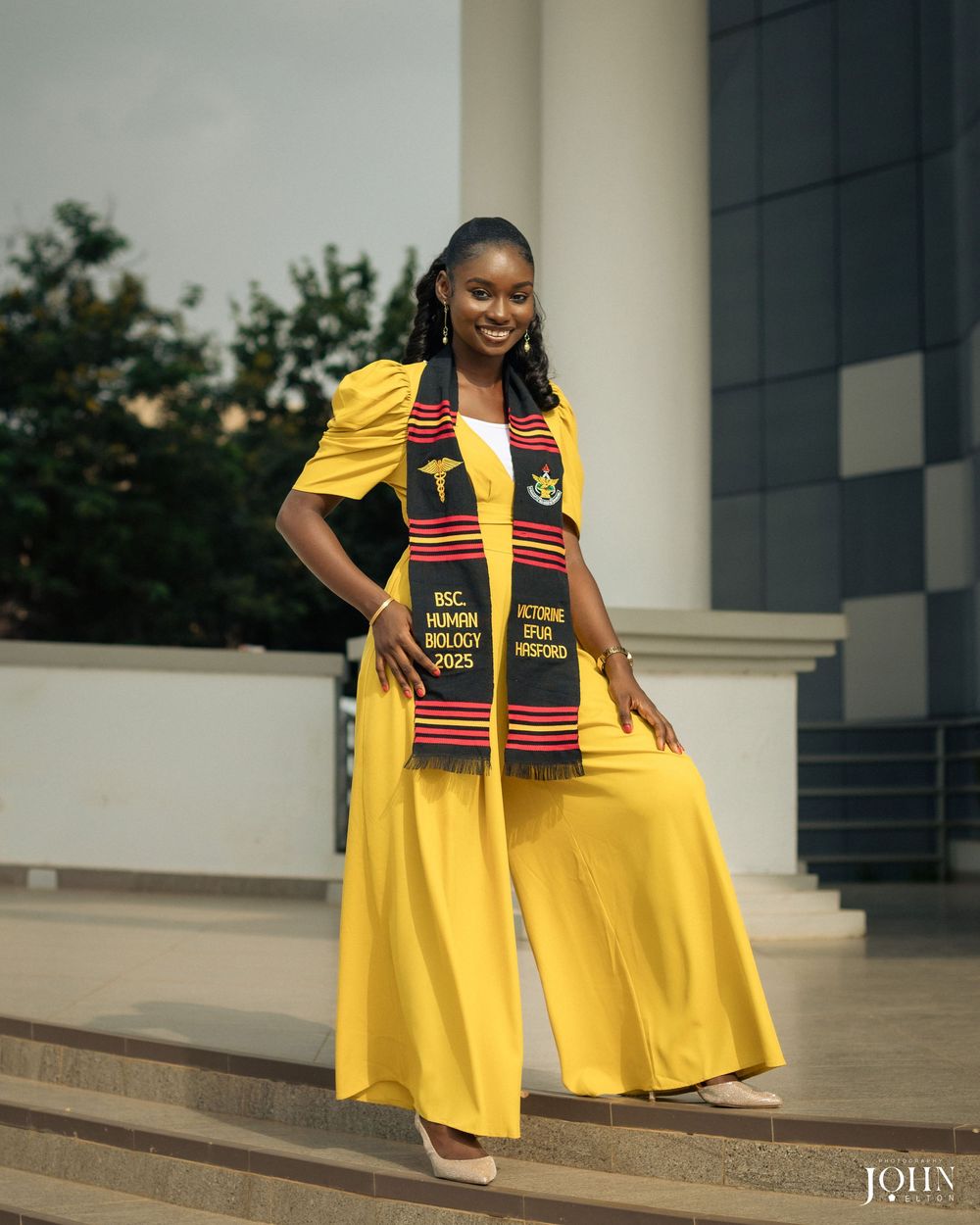 Graduate in yellow academic regalia and colorful kente stole poses for graduation photos on campus.