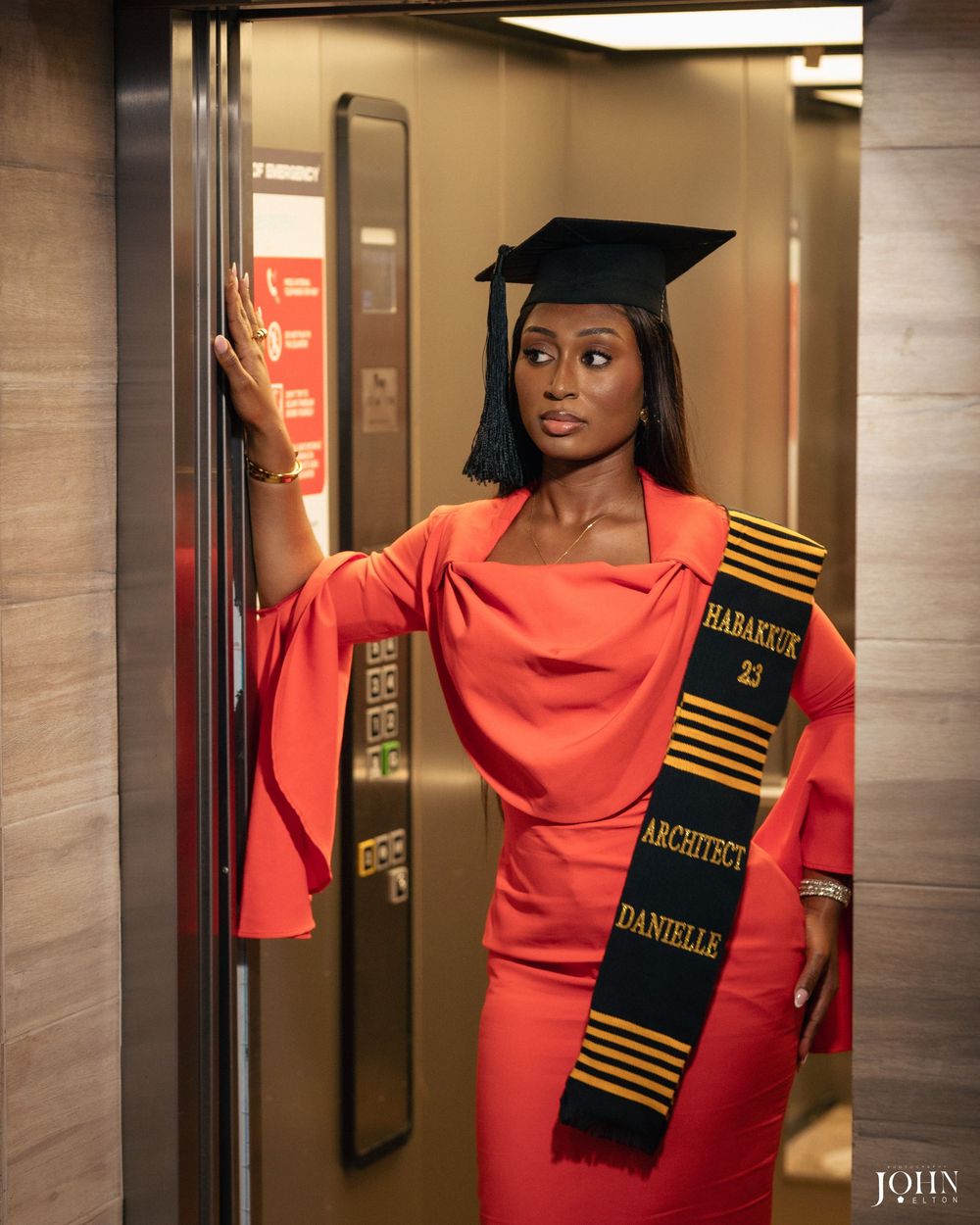 Graduate wearing a bright coral dress and graduation stole poses in front of elevator doors during commencement.