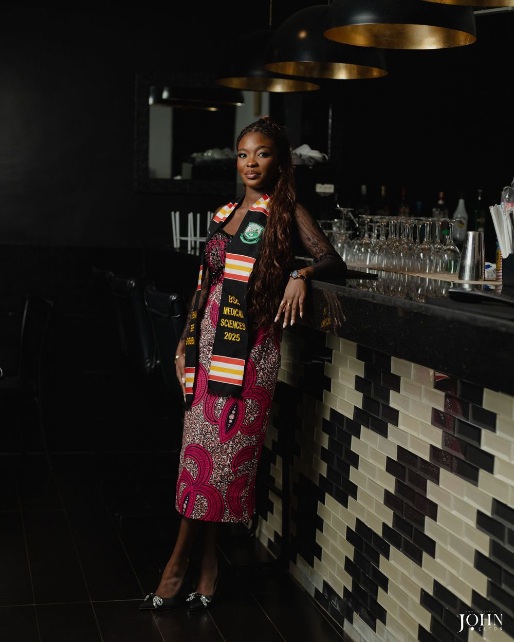 A person wearing a pink and brown patterned dress poses against a geometric black and white wall in dim lighting.