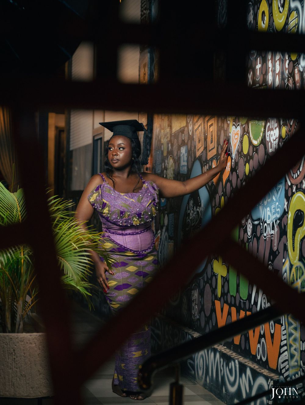 A graduate in purple attire poses artistically against a wall with vibrant murals.