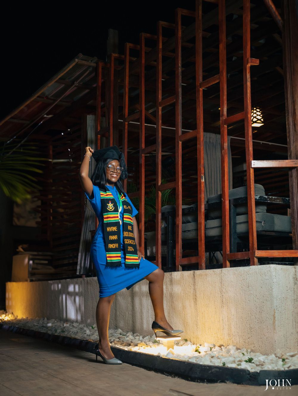 Graduate in blue shirt and graduation stole poses on outdoor terrace with decorative lighting.