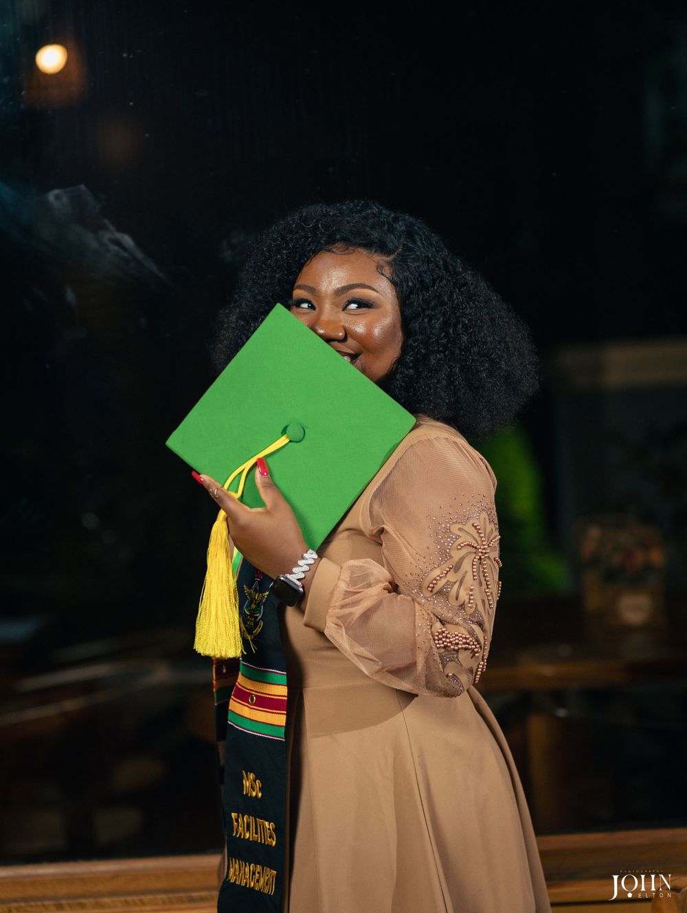 Graduate in tan dress holds green graduation folder during evening ceremony.