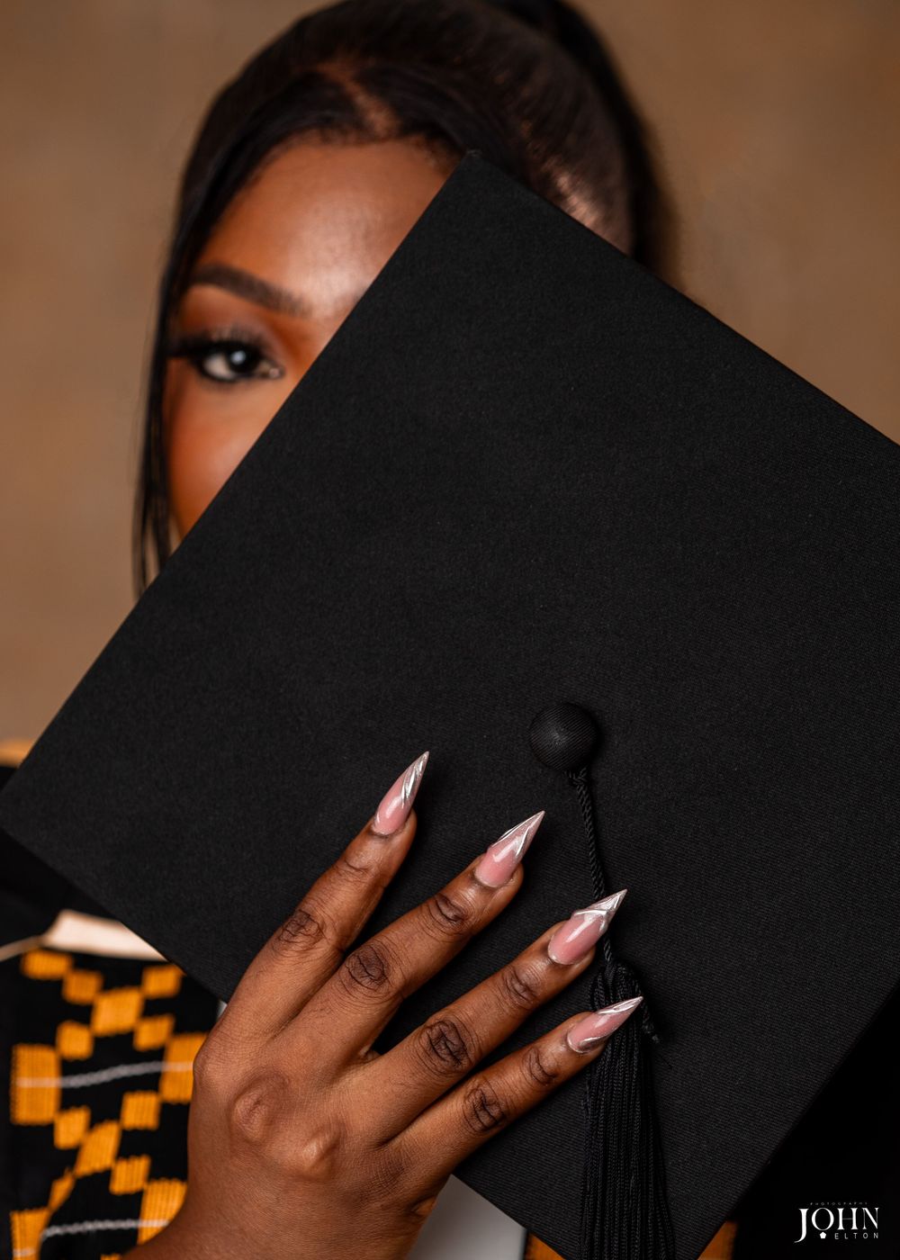 A hand with long stiletto nails holds a black graduation cap against a warm-toned background.