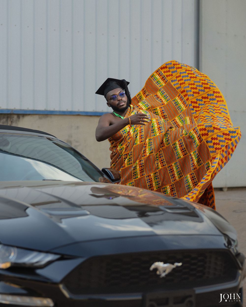 A graduate in academic regalia poses playfully with vibrant kente cloth against a black Mustang car.