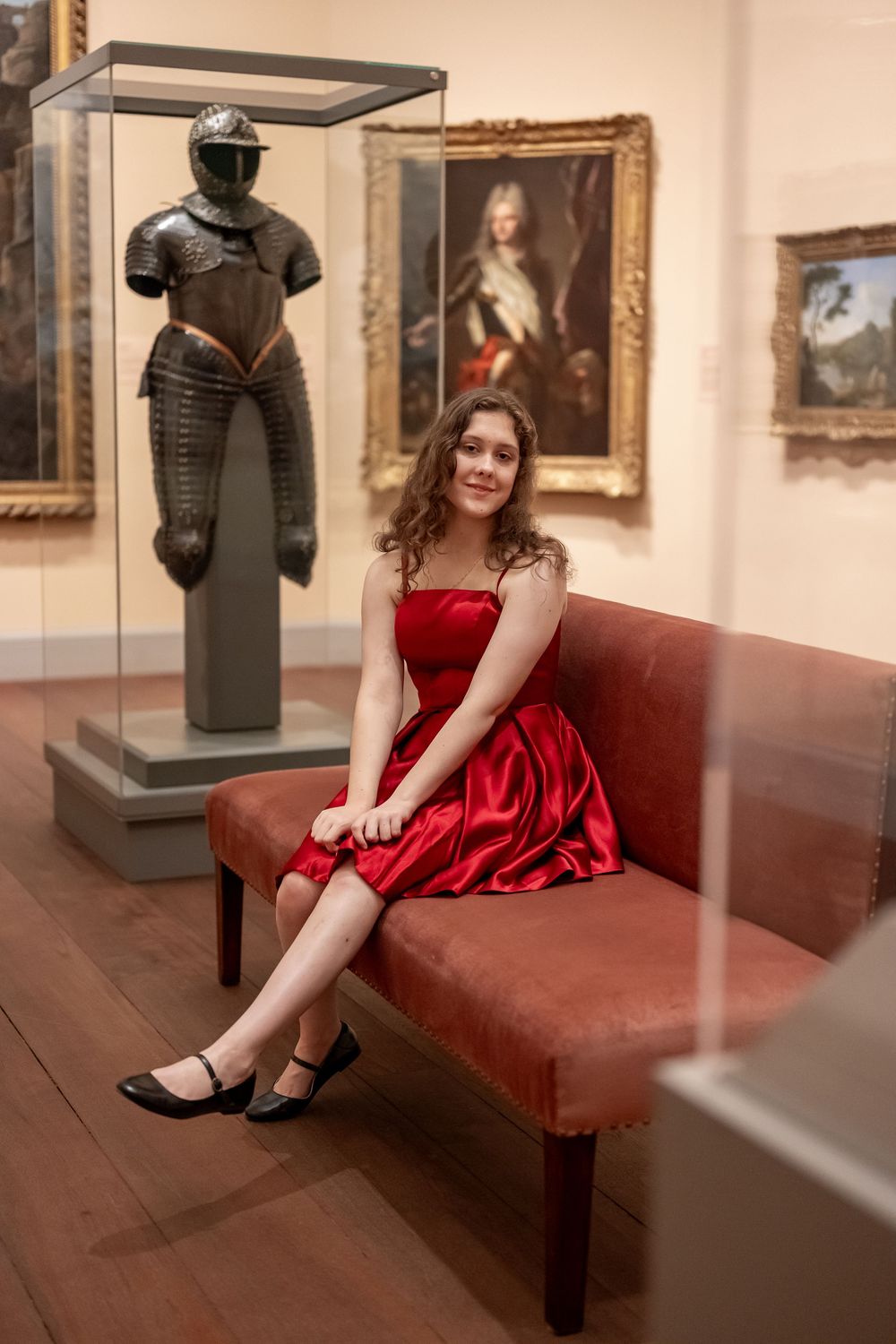 Woman in elegant red dress sits on brown leather bench in art gallery surrounded by paintings and sculptures.