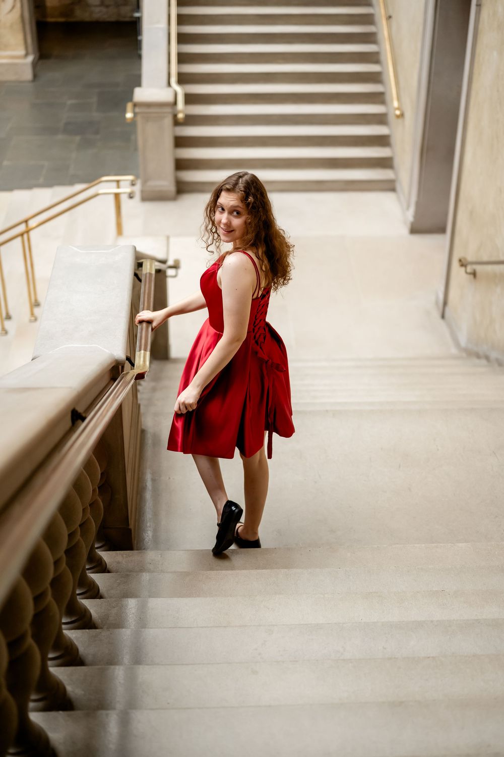 A woman in a red dress poses gracefully on concrete steps in an architectural setting.