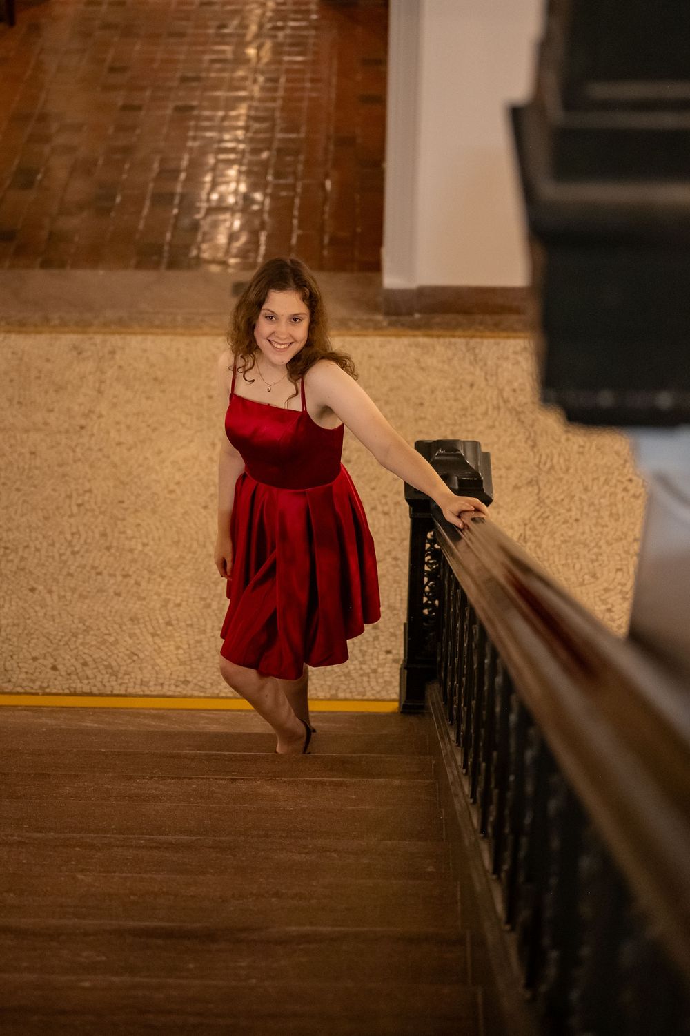 A person in a red velvet dress poses on wooden stairs with an ornate railing in warm lighting.