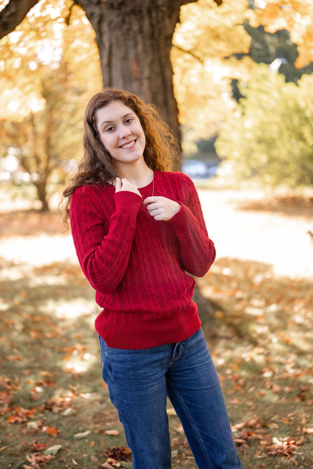 A cozy autumn scene with someone wearing a red cable knit sweater and jeans standing among fallen leaves outdoors.