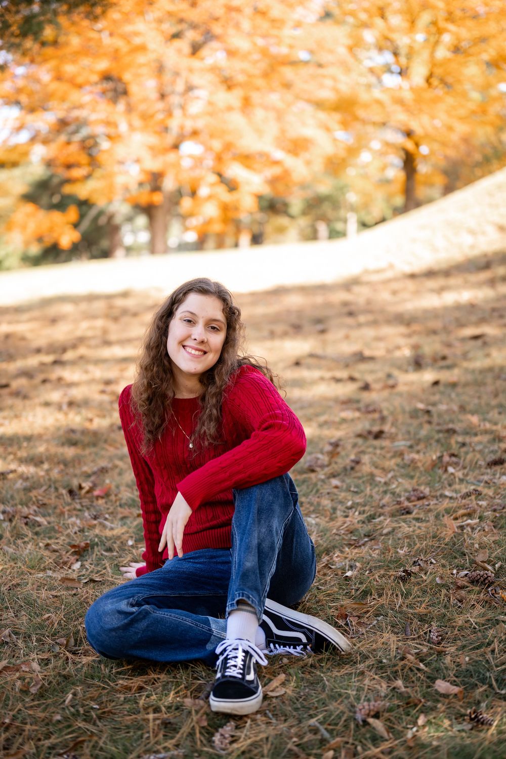 Person in red sweater and jeans sits on autumn ground with orange foliage in background.