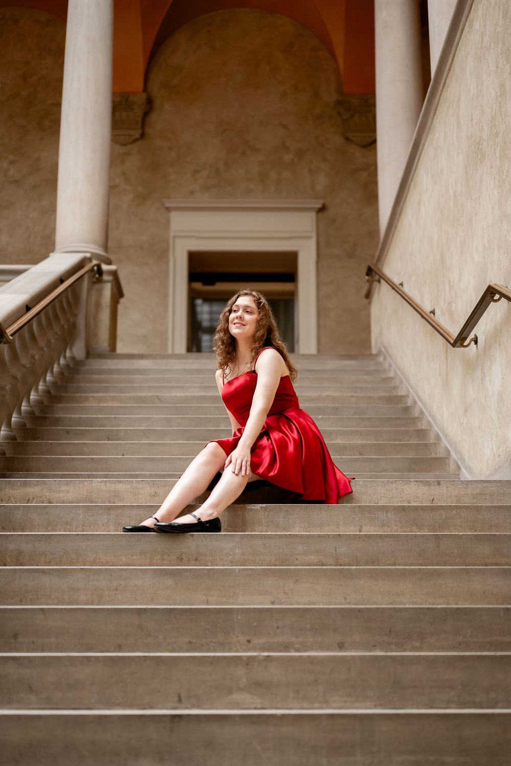 A woman in a red dress sits gracefully on concrete steps of a grand building entrance.