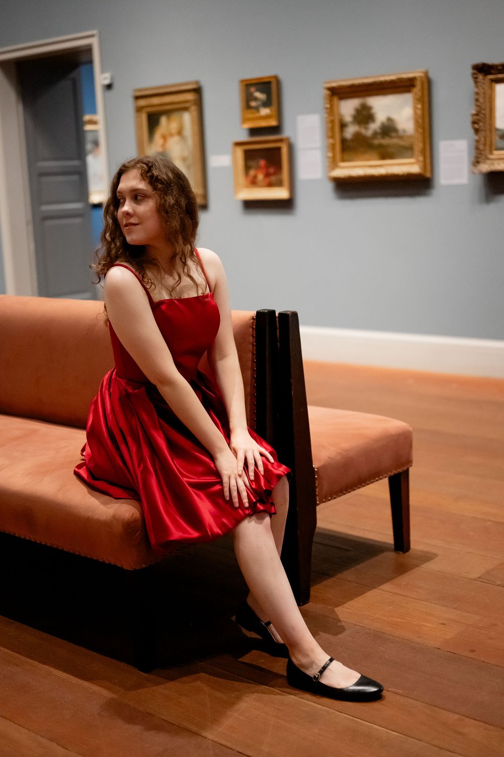 Figure in a red dress sits on a wooden bench in an art gallery with framed paintings displayed on the walls.