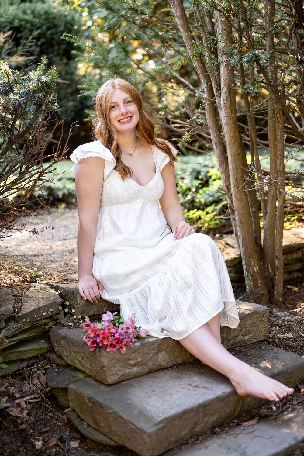 Person in white dress sits on stone steps in garden setting holding pink flowers in outdoor portrait session.