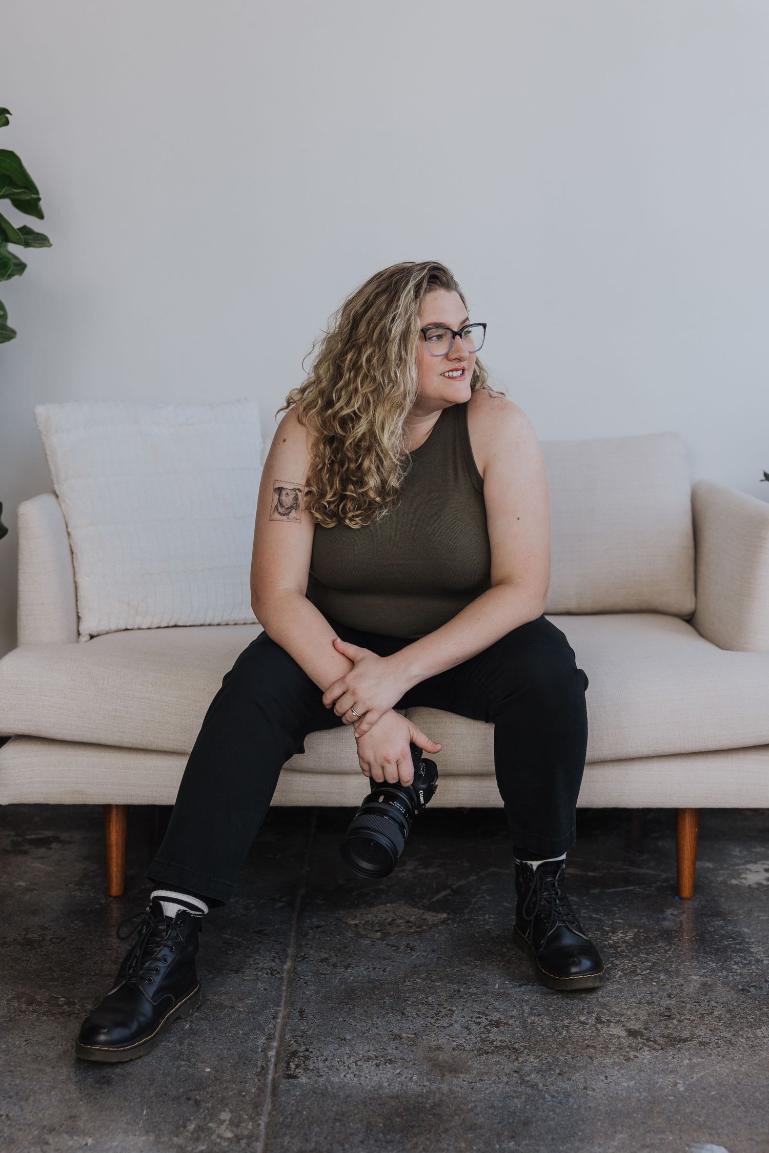 A photographer wearing a brown tank top and dark jeans sits casually on a light colored couch with a plant in the background.