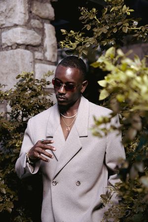 A person in a cream double-breasted blazer poses against a stone wall with foliage in an editorial style photoshoot.