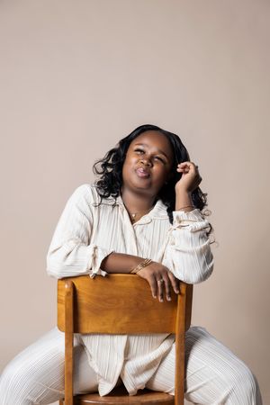 A person in a white dress shirt sits casually on a wooden chair against a neutral beige background.