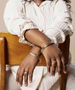 Close-up view of hands wearing white clothing and jewelry resting on wooden chairs.